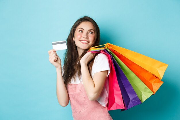 Joven mujer satisfecha sonriendo, mostrando tarjeta de crédito plástica y sosteniendo bolsas de compras, comprando con pago sin contacto, de pie sobre fondo azul.