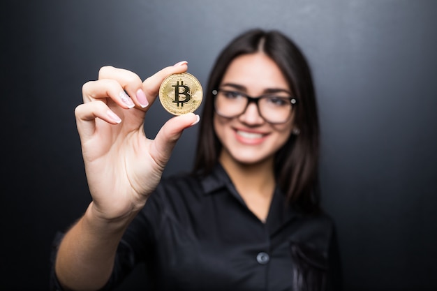 Joven mujer segura de éxito con gafas sostiene un bitcoin de oro en su mano aislado en la pared negra