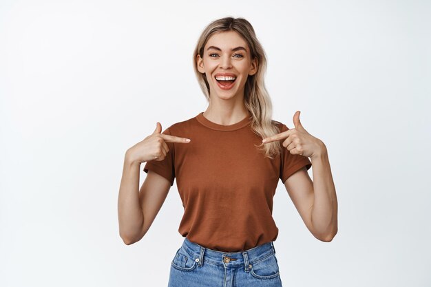 Joven mujer segura de sí misma sonriendo y apuntándose con el dedo a sí misma mostrando una camiseta de pie contra el fondo blanco