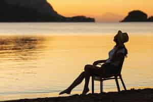 Foto gratuita joven mujer sentada en el asiento en la puesta de sol en la orilla de un lago