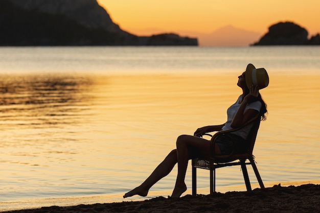 Joven mujer sentada en el asiento en la puesta de sol en la orilla de un lago