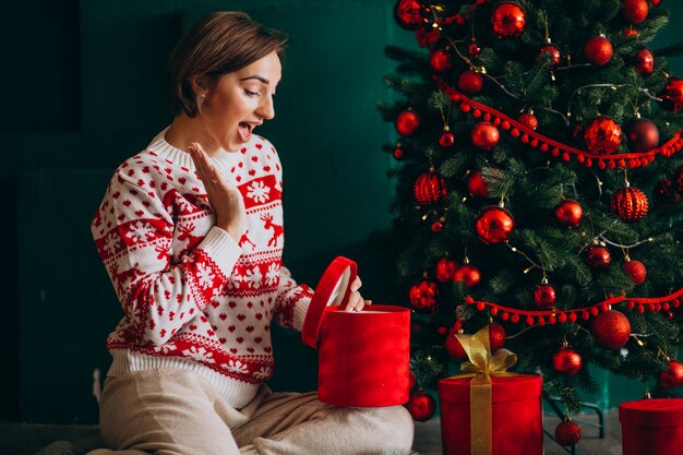 Joven mujer sentada junto al árbol de Navidad con cajas rojas