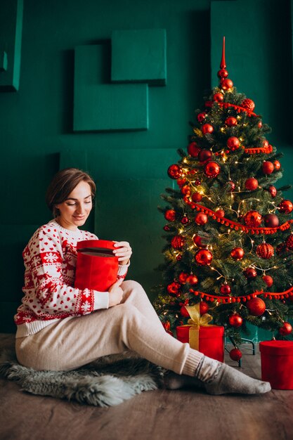 Joven mujer sentada junto al árbol de Navidad con cajas rojas