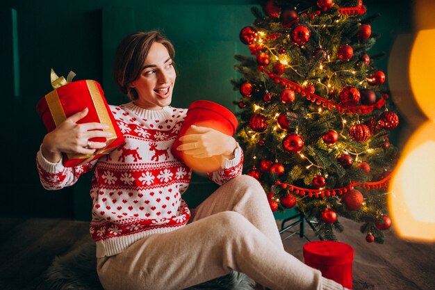 Joven mujer sentada junto al árbol de Navidad con cajas rojas