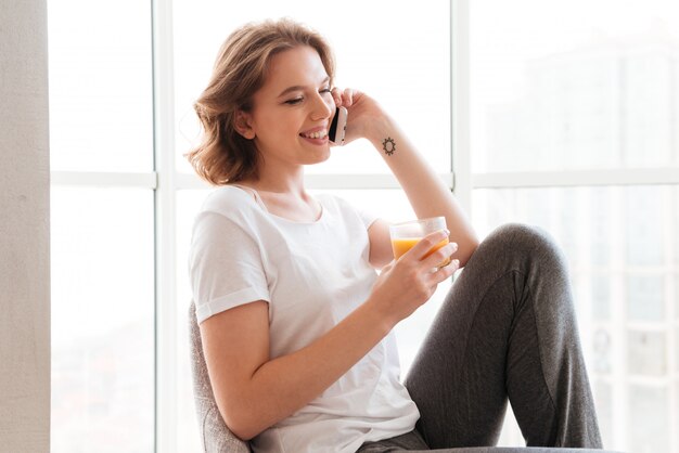 Joven mujer sentada junto a la ventana bebiendo jugo hablando por teléfono