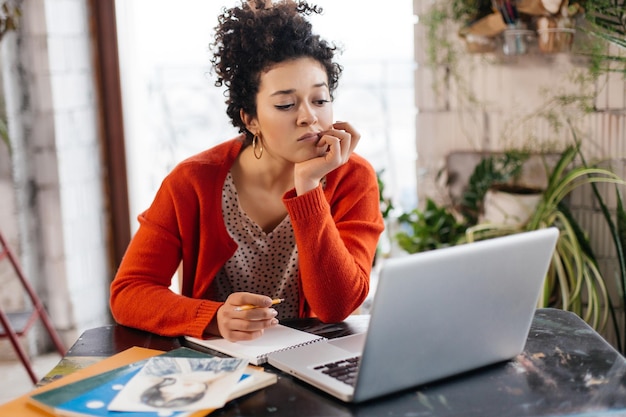 Joven mujer seria con cabello rizado oscuro sentada en la mesa trabajando cuidadosamente en una laptop en un taller moderno y acogedor con grandes ventanas en el fondo