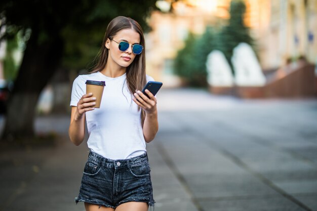 Joven mujer sexy con café para llevar y teléfono caminando en la calle de verano