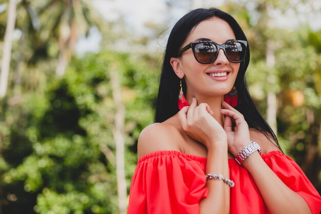 Joven mujer sexy con estilo en vestido rojo de verano de pie en la terraza de un hotel tropical, fondo de palmeras, pelo largo y negro, gafas de sol, pendientes étnicos, gafas de sol, mirando hacia adelante, de cerca