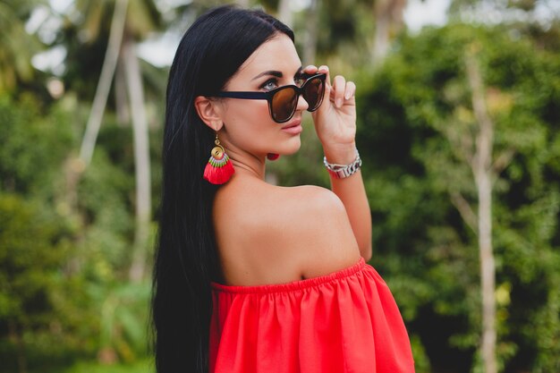 Joven mujer sexy con estilo en vestido rojo de verano de pie en la terraza de un hotel tropical, fondo de palmeras, pelo largo y negro, gafas de sol, pendientes étnicos, gafas de sol, mirando hacia adelante, de cerca