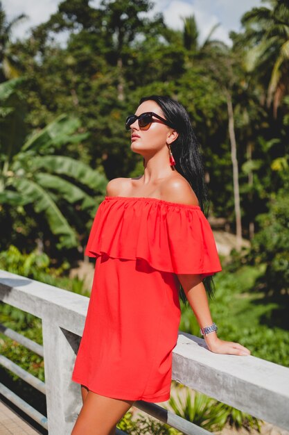 Joven mujer sexy con estilo en vestido rojo de verano de pie en la terraza de un hotel tropical, fondo de palmeras, pelo largo y negro, gafas de sol, pendientes étnicos, gafas de sol, sonriendo