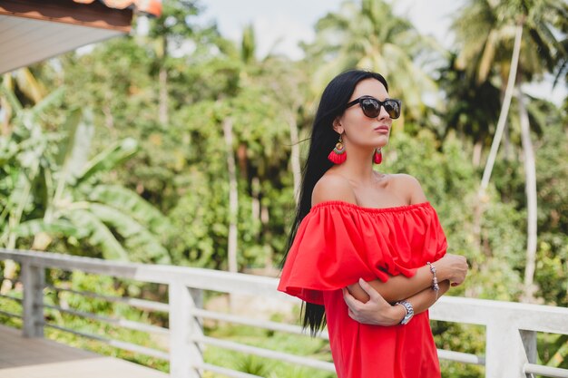 Joven mujer sexy con estilo en vestido rojo de verano de pie en la terraza de un hotel tropical, fondo de palmeras, pelo largo y negro, gafas de sol, pendientes étnicos, gafas de sol, sonriendo