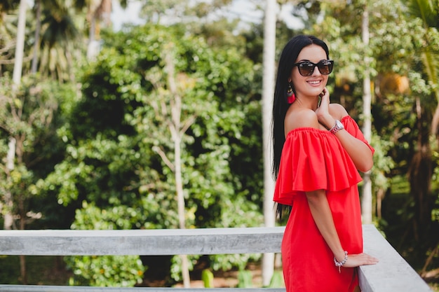 Joven mujer sexy con estilo en vestido rojo de verano de pie en la terraza de un hotel tropical, fondo de palmeras, pelo largo y negro, gafas de sol, pendientes étnicos, gafas de sol, sonriendo