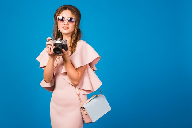 Joven mujer sexy con estilo en vestido rosa de lujo, tendencia de la moda de verano, estilo chic, gafas de sol, tomando fotografías con cámara vintage