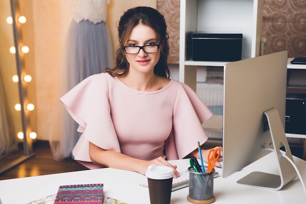 Joven mujer sexy con estilo en vestido rosa de lujo, tendencia de verano, estilo chic, diseñador de moda que trabaja en la oficina en la computadora