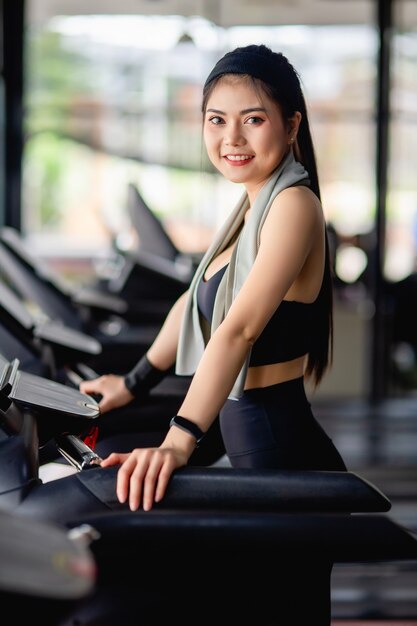 Joven mujer sexy con ropa deportiva, tela a prueba de sudor y reloj inteligente caminando en la cinta de correr calentar antes de correr para entrenar en el gimnasio moderno, sonrisa, espacio de copia