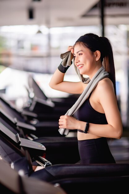 Joven mujer sexy con ropa deportiva, tela a prueba de sudor y reloj inteligente usa una toalla, limpia el sudor en la frente durante el entrenamiento en el gimnasio moderno, sonríe y mira por la ventana, copia espacio