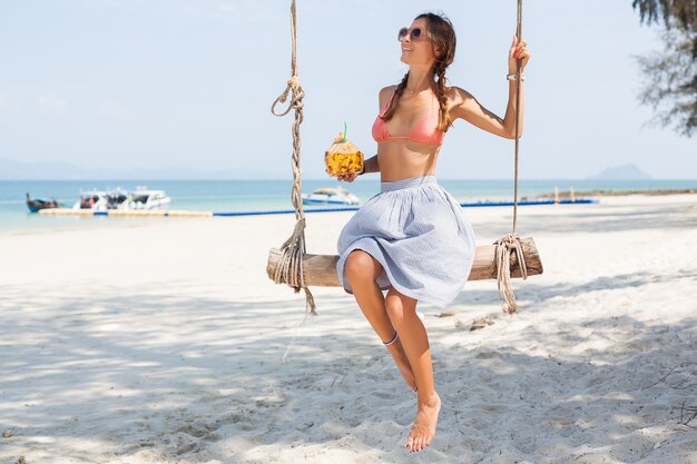 Joven mujer sexy sentada en columpio en la playa tropical, vacaciones de verano, estilo de moda, falda, parte superior del bikini, bebiendo cóctel de coco, sonriendo, relajante