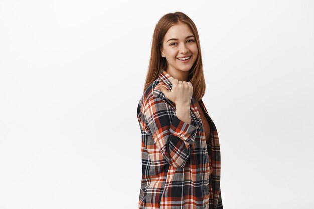 Joven mujer sincera, mujer apuntando con el pulgar a la izquierda detrás de su hombro, sonriendo amistosamente y confiada al frente, mostrando el camino, recomendar el lugar, de pie contra la pared blanca