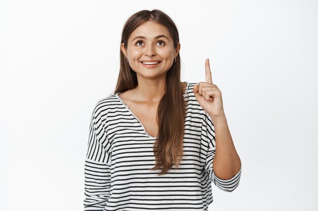 Joven mujer sonriente apuntando, mirando hacia arriba con expresión de cara feliz en blanco.