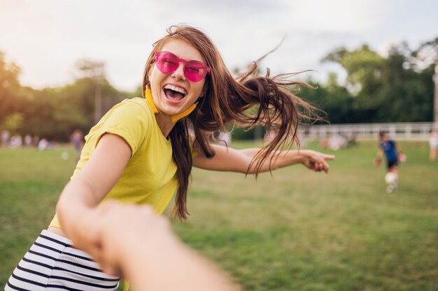 Joven mujer sonriente bastante elegante divirtiéndose en el parque de la ciudad, de la mano de su novio, sígueme, positivo emocional, con top amarillo, gafas de sol rosas, tendencia de moda de estilo veraniego