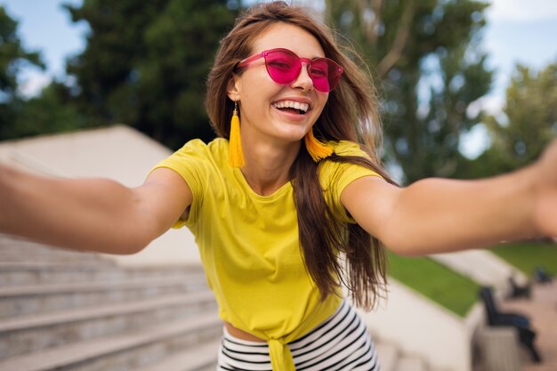 Joven mujer sonriente bastante elegante haciendo foto selfie en el parque de la ciudad, positiva, emocional, con top amarillo, gafas de sol rosas, tendencia de moda de estilo veraniego, cabello largo, divirtiéndose