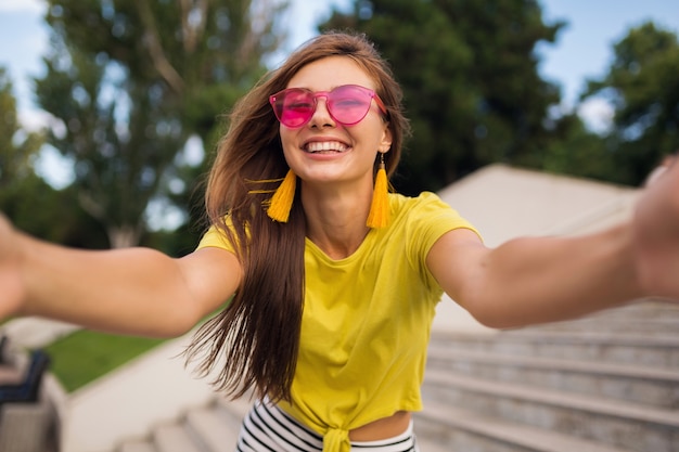 Joven mujer sonriente bastante elegante haciendo selfie en el parque de la ciudad, positiva, emocional, con top amarillo, gafas de sol rosa, tendencia de moda de estilo veraniego, cabello largo, divirtiéndose