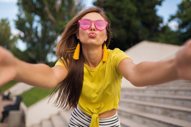 Joven mujer sonriente bastante elegante haciendo selfie en el parque de la ciudad, positiva, emocional, con top amarillo, gafas de sol rosa, tendencia de moda de estilo veraniego, cabello largo, divirtiéndose