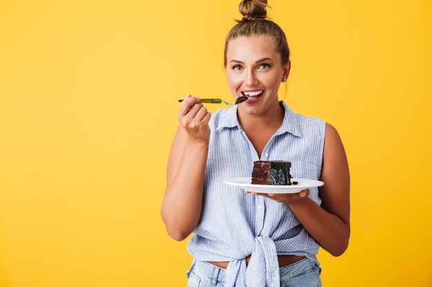 Joven mujer sonriente en camisa mirando felizmente a la cámara mientras come pastel de chocolate sobre fondo amarillo