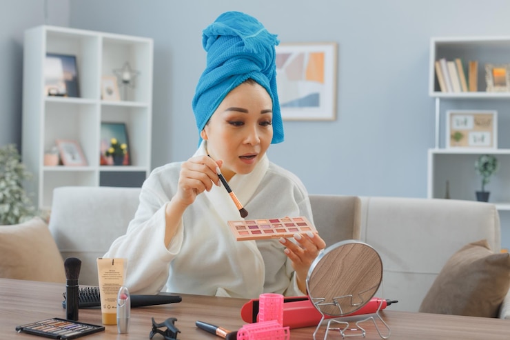 Foto gratuito joven mujer sonriente feliz asiática con una toalla en la cabeza sentada en el tocador en el interior de la casa aplicando sombras de ojos mirando al espejo haciendo la rutina de maquillaje matutino