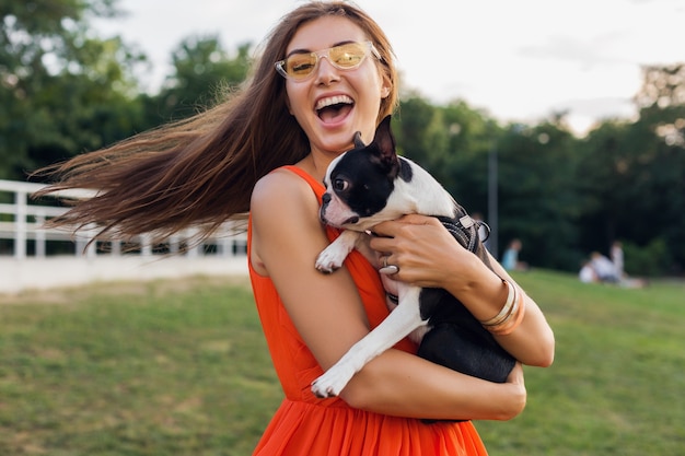 Joven mujer sonriente feliz con perro boston terrrier en el parque, día soleado de verano, estado de ánimo alegre, jugando con la mascota, agitando el pelo largo, divirtiéndose, con gafas de sol, riendo