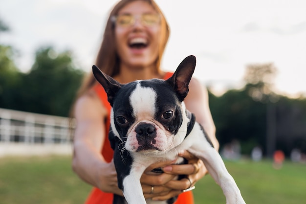 Foto gratuita joven mujer sonriente feliz con perro boston terrrier en el parque, día soleado de verano, estado de ánimo alegre, jugando con la mascota, agitando el pelo largo, divirtiéndose, tendencia de la moda de verano