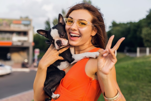 Joven mujer sonriente feliz con perro boston terrrier en el parque, día soleado de verano, estado de ánimo alegre, jugando con la mascota, agitando el pelo largo, divirtiéndose, tendencia de la moda de verano