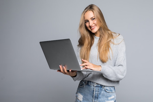 Joven mujer sonriente feliz en ropa casual sosteniendo una computadora portátil y enviando un correo electrónico a su mejor amiga aislada sobre fondo gris