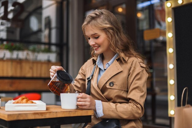 Joven mujer sonriente con gabardina sosteniendo felizmente una tetera y una taza blanca al aire libre en la acogedora terraza del café