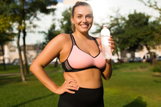 Joven mujer sonriente de talla grande con top deportivo rosa y calzas mirando alegremente a la cámara con una botella de agua pura en la mano mientras pasa tiempo en el parque de la ciudad