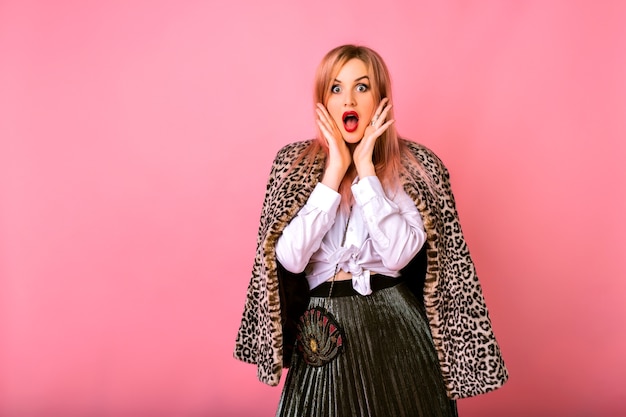 Joven mujer sorprendida sorprendida divertida posando sobre fondo rosa, vestida con camisa blanca y abrigo de leopardo, emociones poderosas.