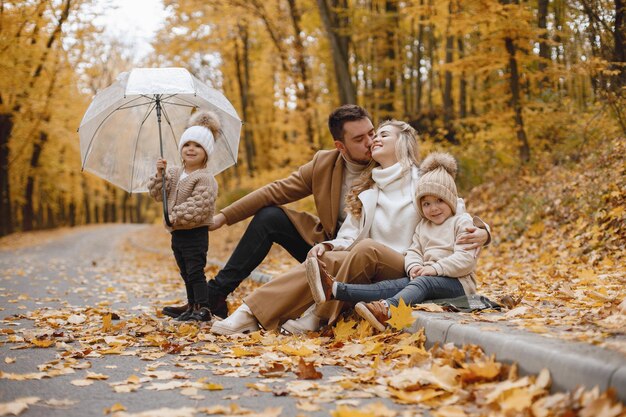 Joven, mujer y sus hijos sentados afuera en el bosque de otoño. Madre rubia y padre moreno abrazándose. Niña sosteniendo un paraguas transparente.