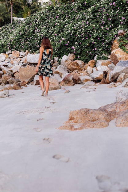 Joven mujer tatuada tranquila en vestido corto con estampado tropical de verano en la playa rocosa con arbusto verde y flores de color rosa púrpura