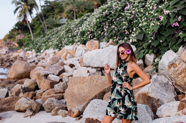 Joven mujer tatuada tranquila en vestido corto con estampado tropical de verano en la playa rocosa con arbusto verde y flores de color rosa púrpura