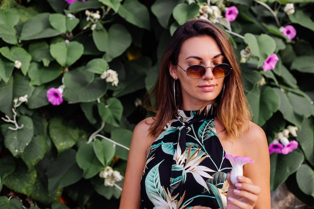 Joven mujer tatuada tranquila en vestido corto con estampado tropical de verano en la playa rocosa con arbusto verde y flores de color rosa púrpura