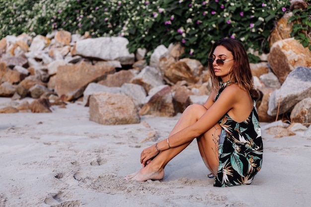 Joven mujer tatuada tranquila en vestido corto con estampado tropical de verano en la playa rocosa con arbusto verde y flores de color rosa púrpura