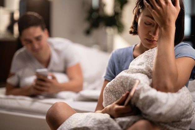 Joven mujer triste con dolor de cabeza sentada en el dormitorio mientras su novio envía mensajes de texto en el teléfono móvil en el fondo