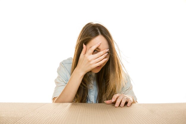 Joven mujer triste en el paquete postal más grande aislado en blanco. Conmocionado modelo femenino molesto en la parte superior de la caja de cartón mirando hacia adentro.