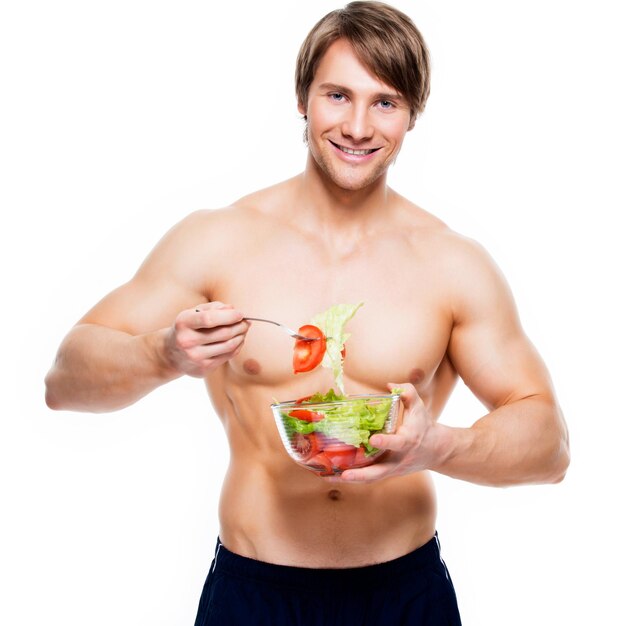 Joven musculoso feliz comiendo una ensalada sobre la pared blanca.