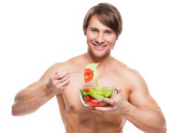 Joven musculoso feliz comiendo una ensalada sobre la pared blanca.