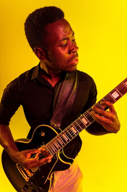 Joven músico afroamericano tocando la guitarra como una estrella de rock en la pared amarilla con luz de neón.