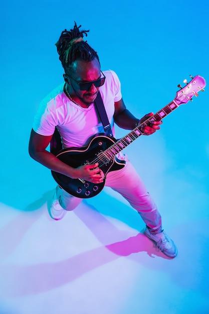 Joven músico afroamericano tocando la guitarra como una estrella de rock en la pared azul con luz de neón.