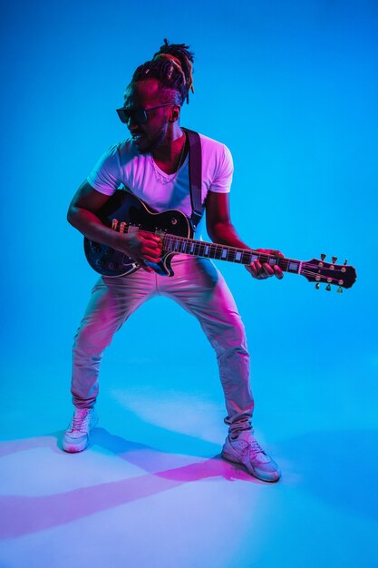 Joven músico afroamericano tocando la guitarra como una estrella de rock sobre fondo azul de estudio en luz de neón. Concepto de música, afición. Chico alegre improvisando. Retrato colorido retro.