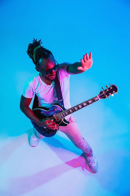 Joven músico afroamericano tocando la guitarra como una estrella de rock sobre fondo azul en luz de neón.