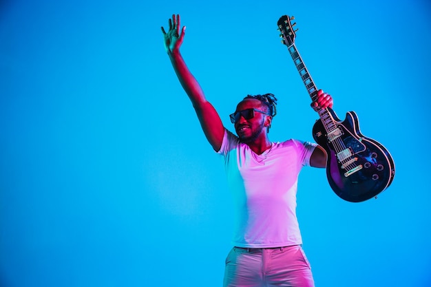 Joven músico afroamericano tocando la guitarra como una estrella de rock sobre fondo azul en luz de neón.